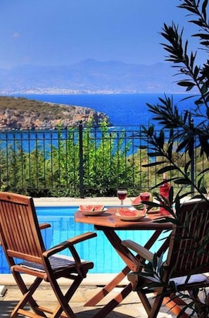 A poolside setting with a view of the sea and mountains, a table with drinks and a watermelon, and two wooden chairs.