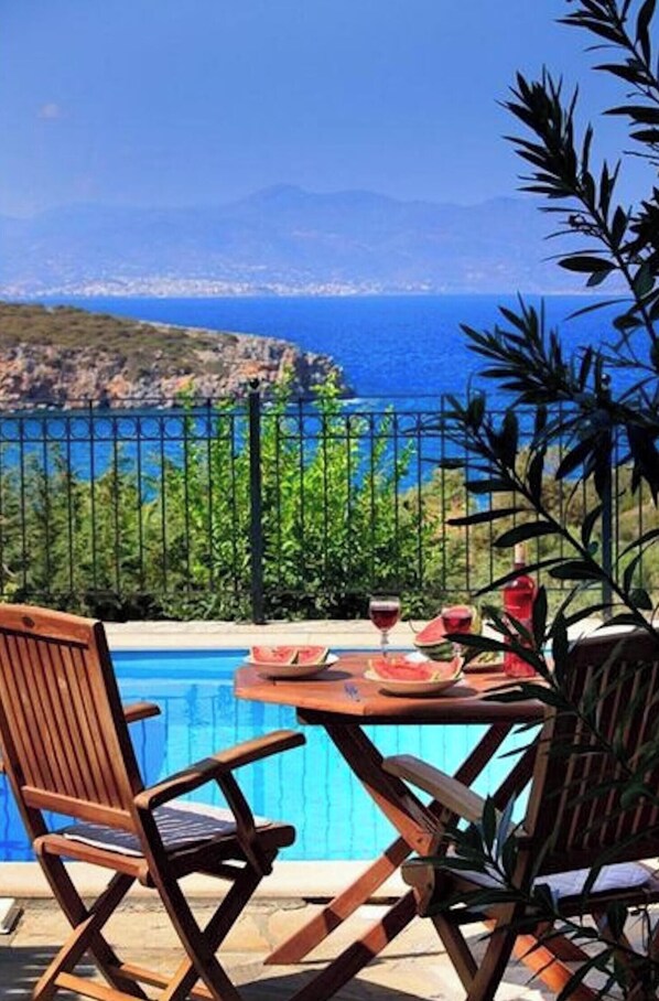 A poolside setting with a view of the sea and mountains, a table with drinks and a watermelon, and two wooden chairs.