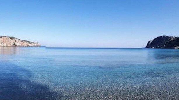 A coastal beach with clear water, rocky cliffs, and a calm sea.
