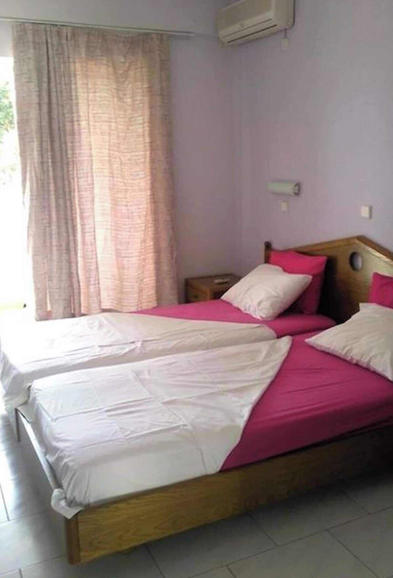 Una cama doble con ropa de cama rosa y blanca, un cabecero de madera, una mesita de noche y una ventana con cortinas.