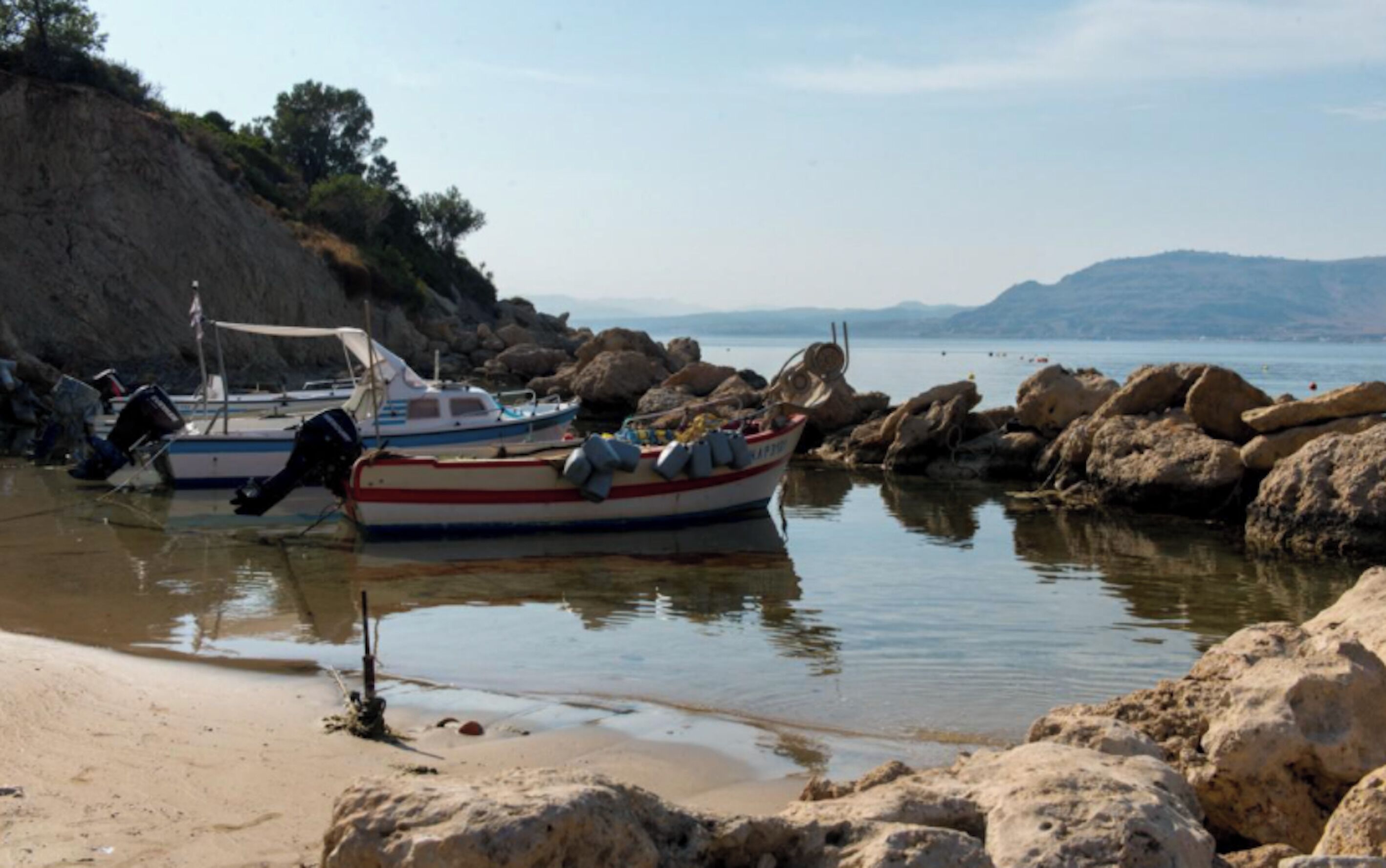 Una barca ormeggiata su una riva rocciosa, con acque calme e colline in lontananza.
