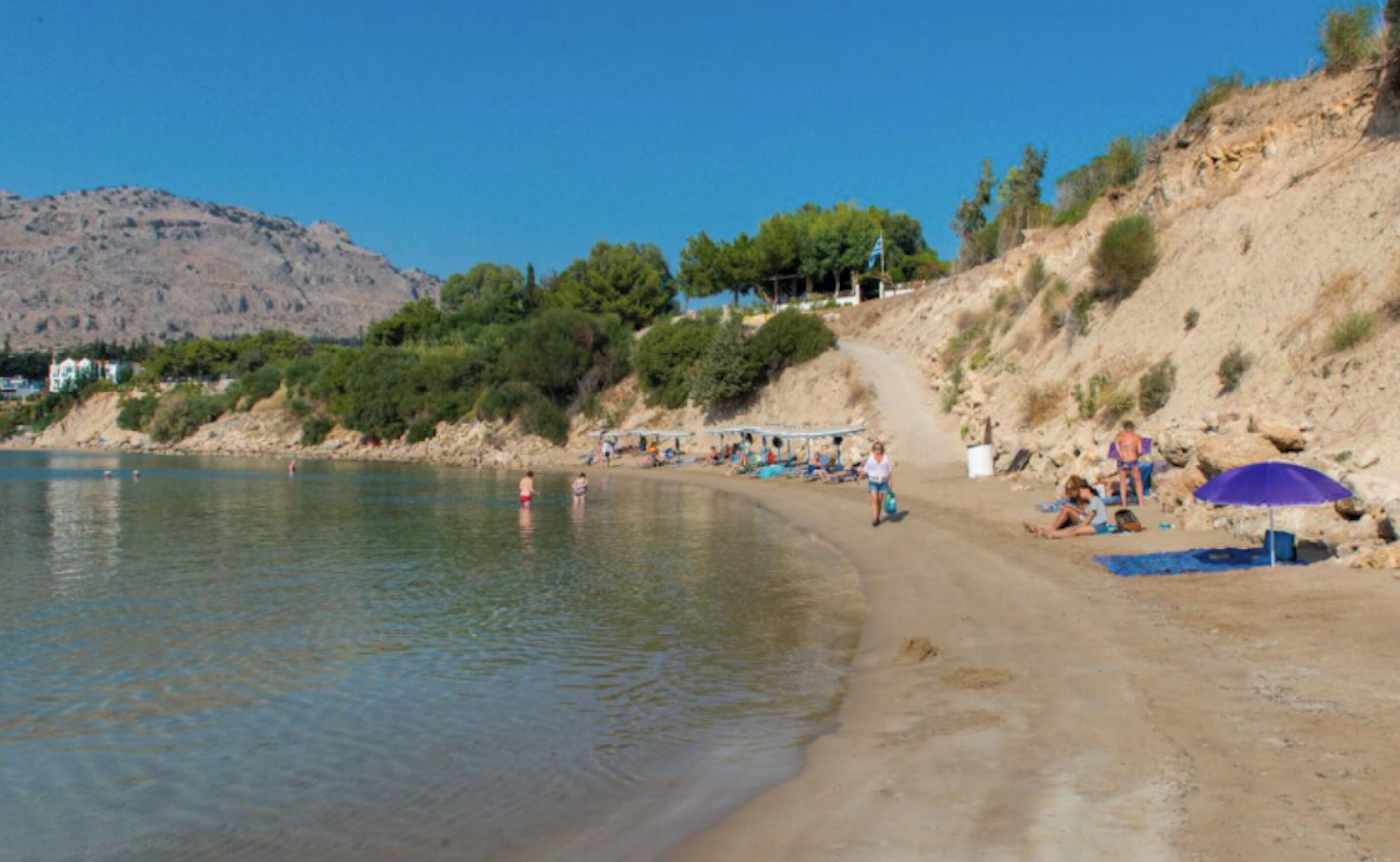 A beach with people sunbathing and swimming, a sandy shore, and a mountain in the background.