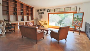 Un vaste salon agrémenté d’une grande bibliothèque en bois, d’un confortable ensemble de canapés, d’une table à manger et d’une grande fenêtre donnant sur un paysage verdoyant.