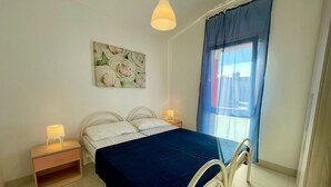 A bedroom with a bed, two bedside tables, a nightstand, a wall-mounted lamp, a window with curtains, and a floral painting on the wall. - Relax in our Flat Near the Coast (Caorle)