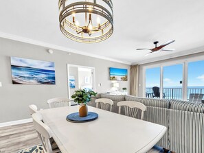 A dining area with a chandelier, a view of the ocean, and a balcony with chairs. - 🌞Pool + Grilling Area-Grandview East 302-Sandscape🌞 (Panama City Beach)