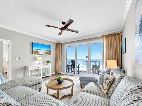 A spacious living room with a large window offering a view of the beach, a ceiling fan, and a cozy seating area with striped sofas and a round coffee table. - 🌞Pool + Grilling Area-Grandview East 302-Sandscape🌞 (Panama City Beach)