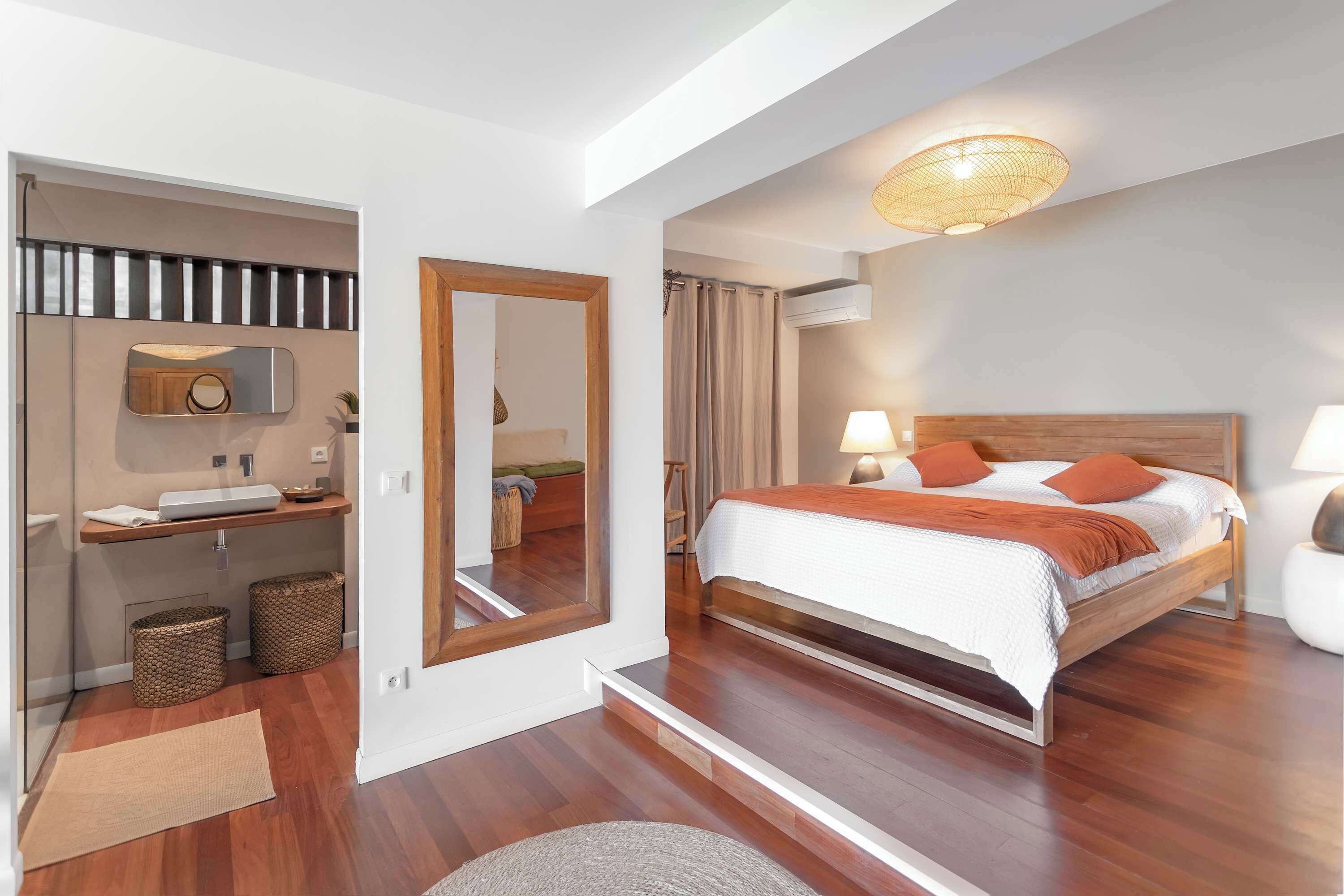 Un dormitorio moderno con cama de madera, una mesita de noche, un espejo, una lámpara colgante y una estantería empotrada con lavamanos y cestas de almacenamiento.