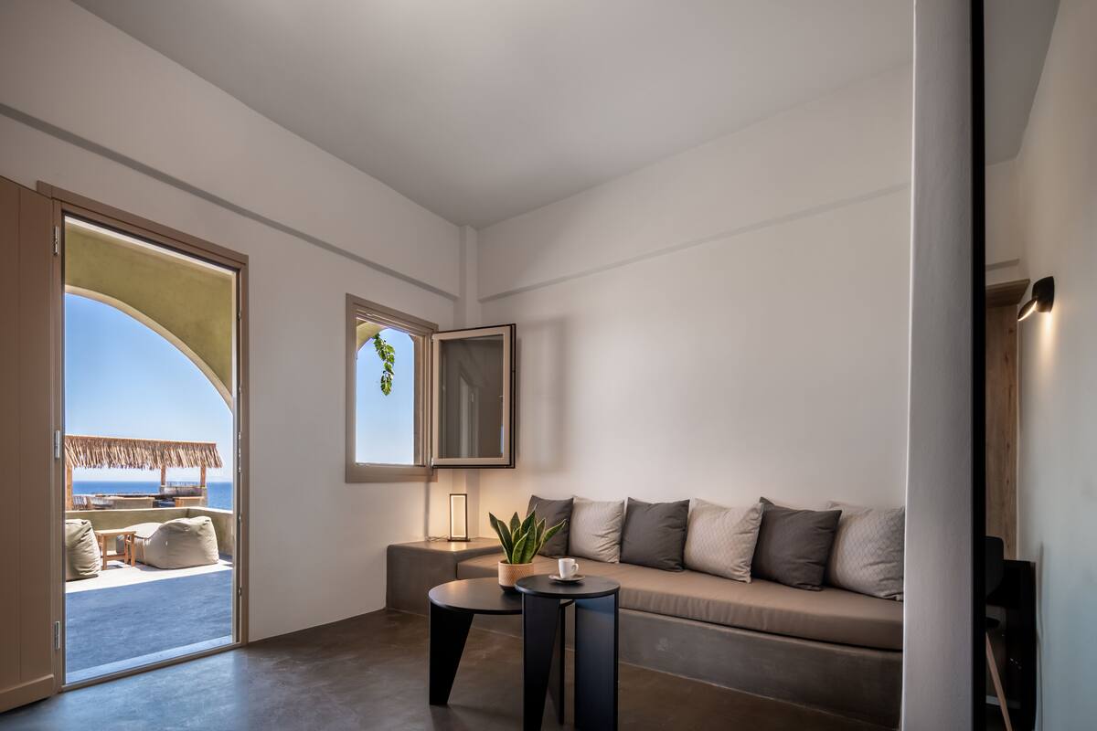 A modern living room with a sofa, two small tables, a potted plant, and a view of the ocean through an open door.