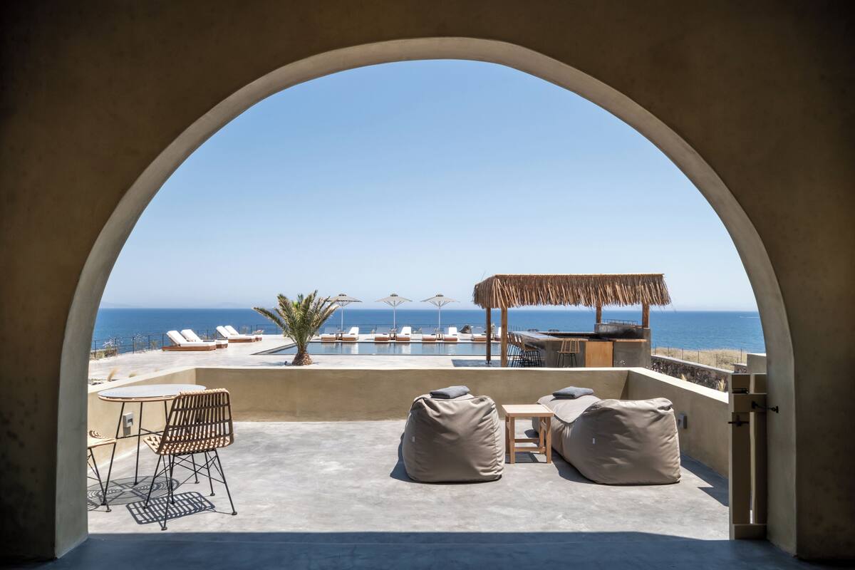 A rooftop terrace with a view of the sea, lounge chairs, a table, and a thatched umbrella.