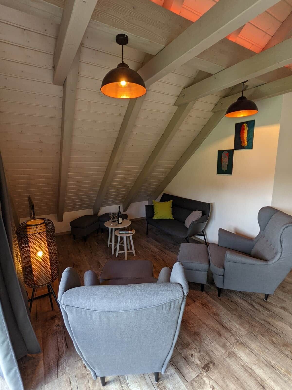A loft-style room with exposed wooden beams, a sofa set, a dining table with chairs, and pendant lighting.