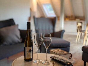 A bottle of wine, two wine glasses, a remote control, and a book on a round table in a modern living room with a sofa and a chair in the background.