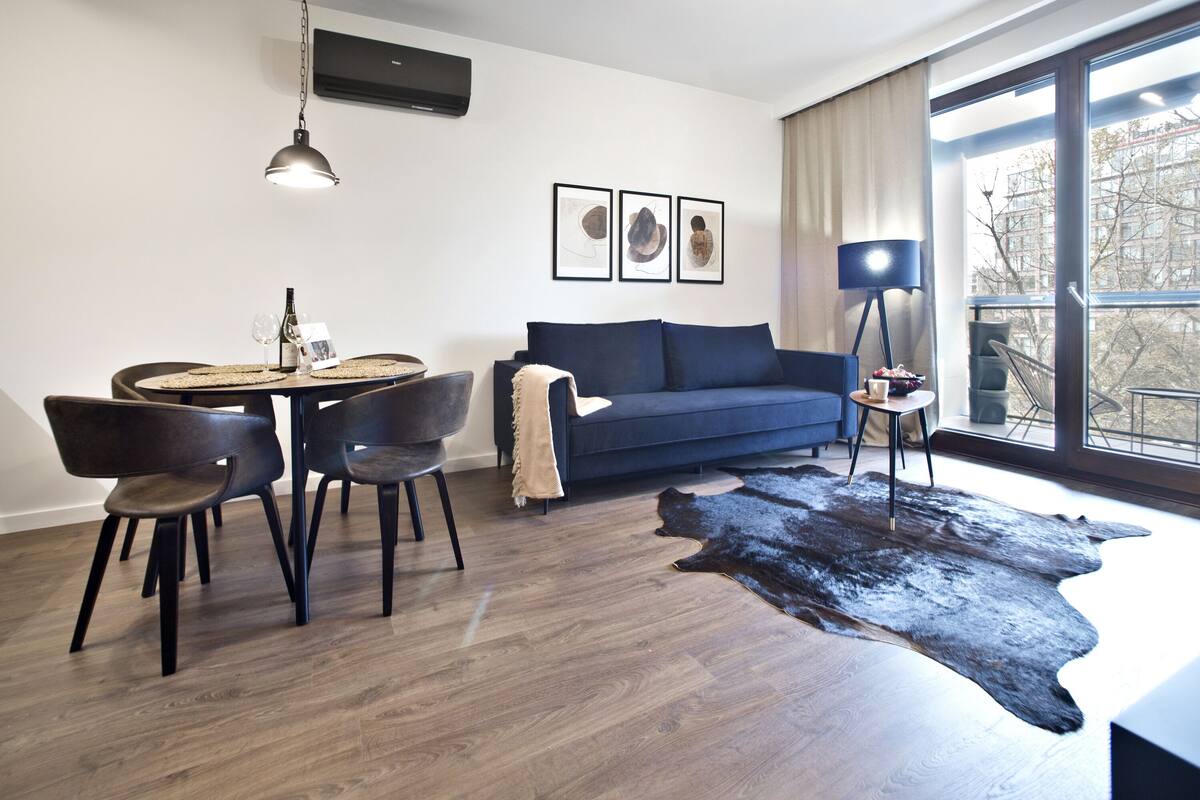 Una sala moderna con un sofá azul oscuro, una mesa de comedor redonda con sillas, un tapete de cuero negro y un ventanal con vistas a árboles.