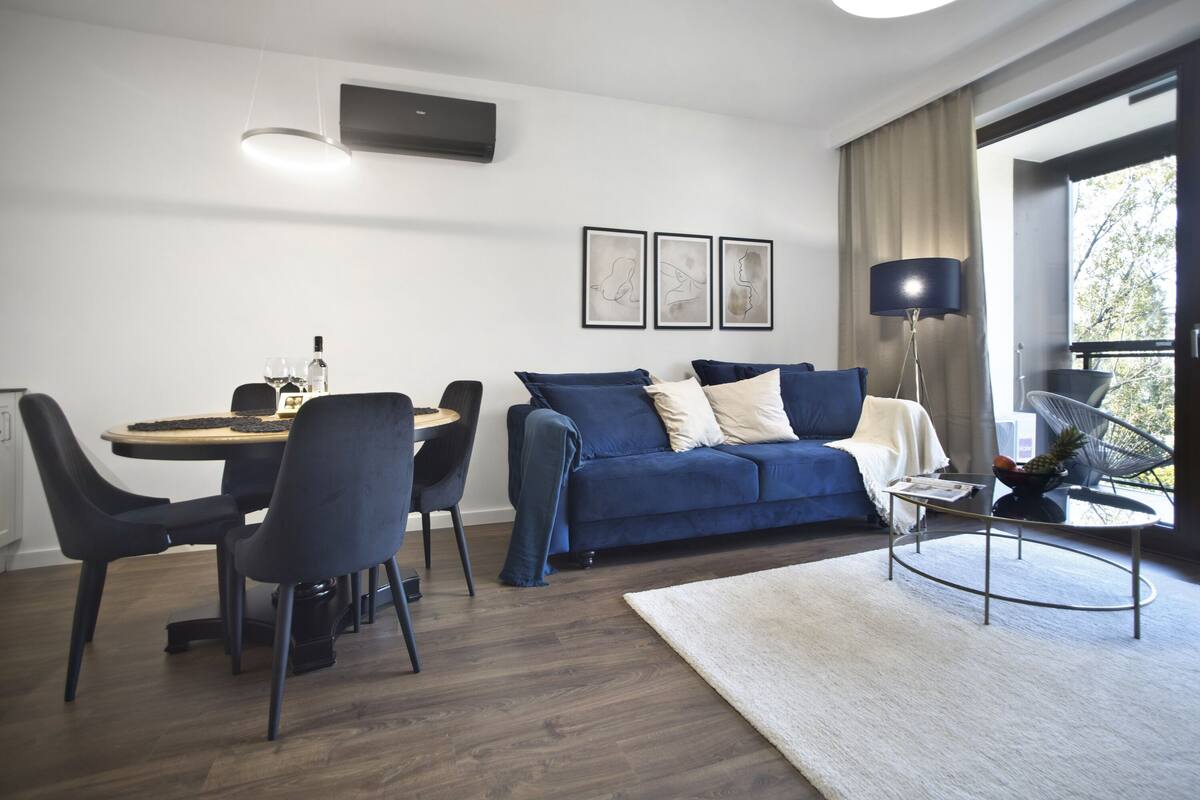Una sala de estar moderna con un sofá azul oscuro, una mesa de comedor redonda con sillas, una mesa de centro y un ventanal con vistas.