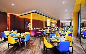 A restaurant with multiple dining tables and chairs, a central bar area, and a television screen on the wall.