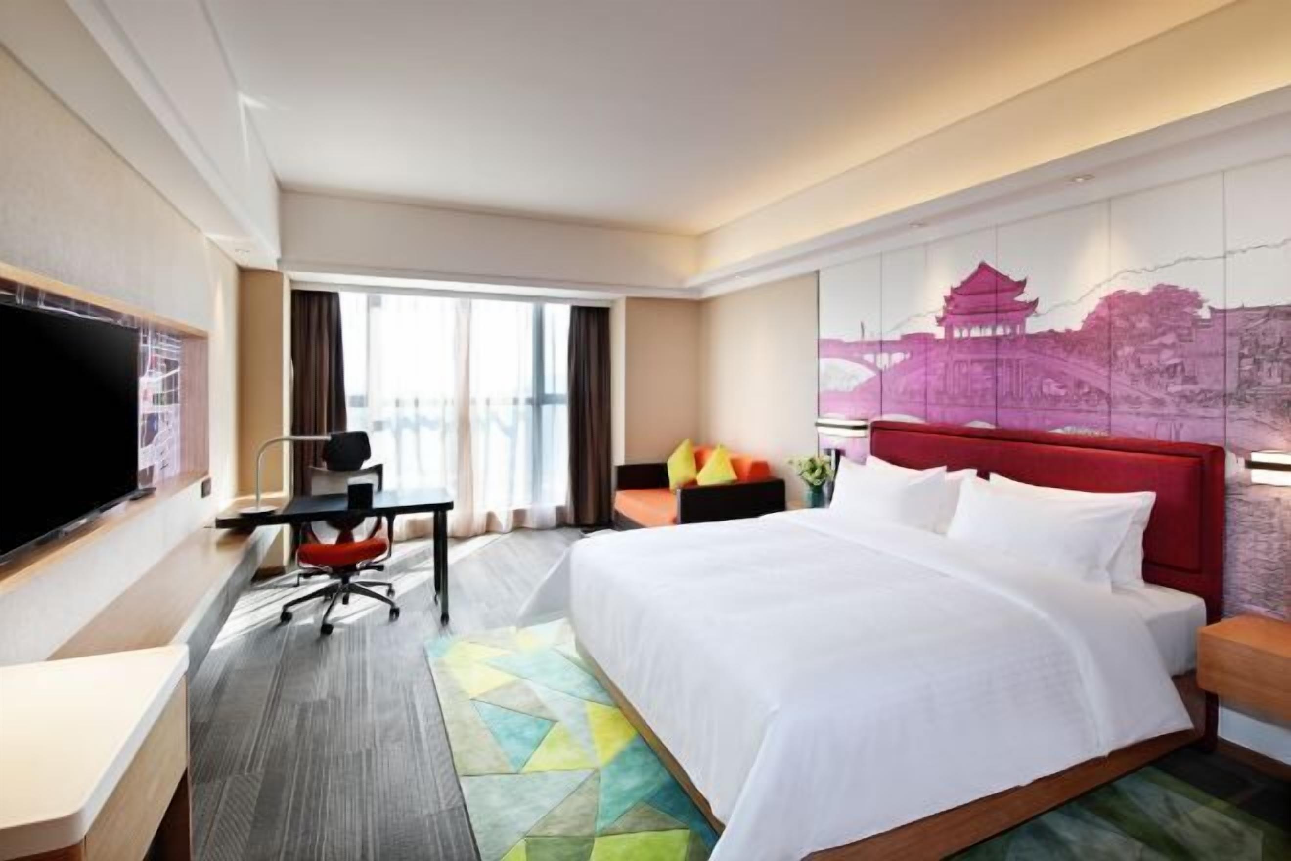 Habitación de hotel con una cama grande, un escritorio con silla, un televisor de pantalla plana y un ventanal con cortinas.