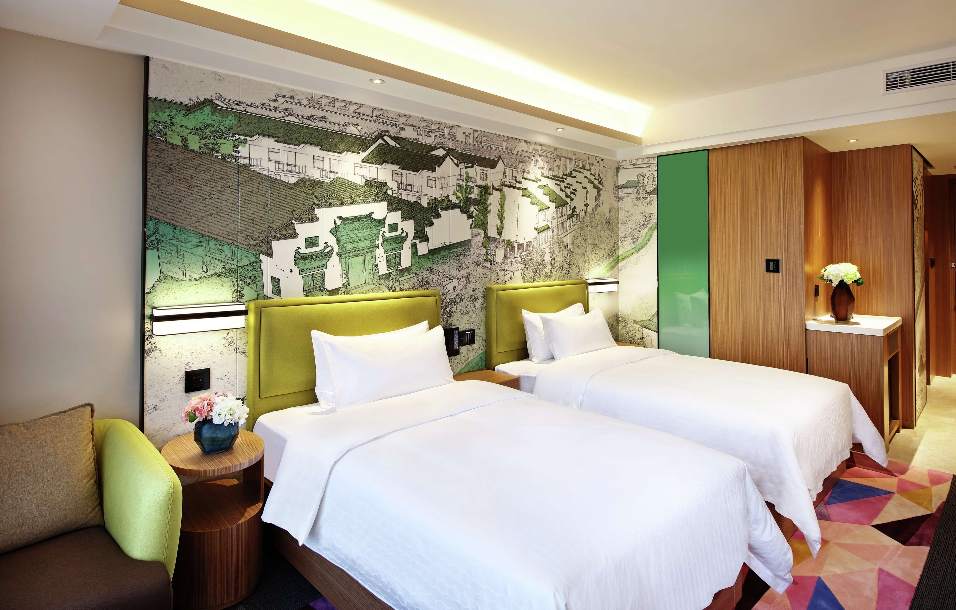 A hotel room with two beds, a green armchair, a small table with a vase, and a mural on the wall.