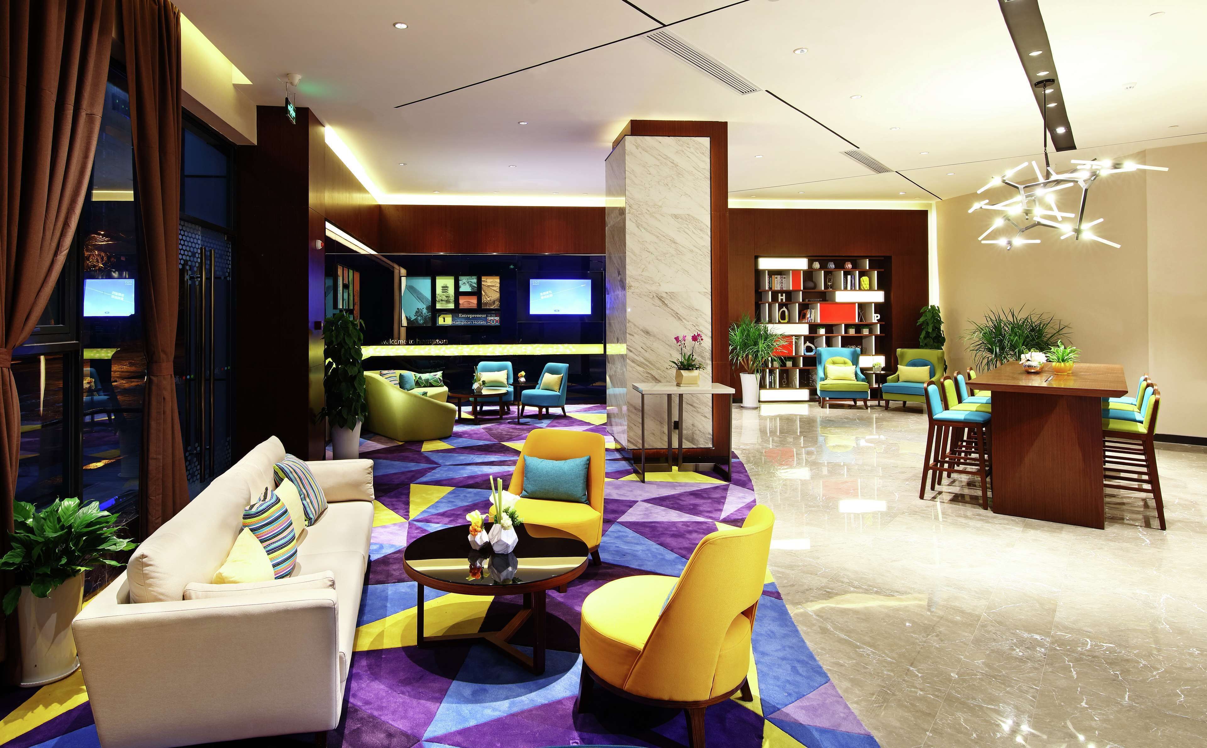 A modern hotel lobby with a seating area, a reception desk, and a television displaying the hotel's name.
