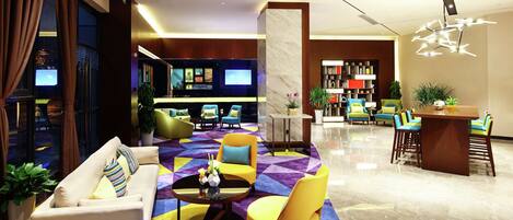 A modern hotel lobby with a seating area, a reception desk, and a television displaying the hotel's name.