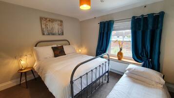 A bedroom with a bed, bedside tables, a window with curtains, a wall-mounted painting, and a vase with flowers.