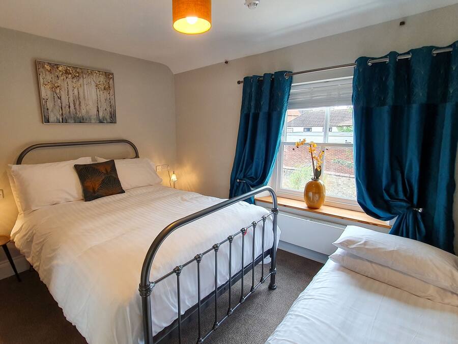 Ein Schlafzimmer mit Bett, Nachttischen, einem Fenster mit Vorhängen, einem Wandbild und einer Vase mit Blumen.