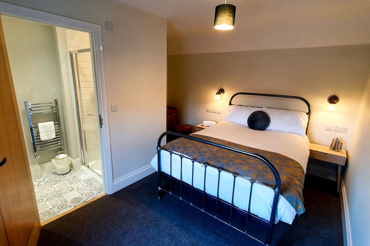 Hotellihuone, jossa on sänky, yöpöytä, lamppu, suihkutila ja ovi.