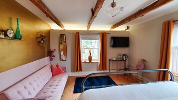 Quarto com sofá rosa, vigas de madeira, televisão, janela com cortinas e mesa de cabeceira com um vaso.