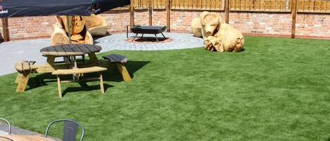 A backyard with artificial grass, a wooden table, chairs, a barbecue grill, and a large umbrella.