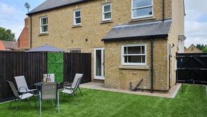 A two-story brick house with a small front yard, a wooden fence, and a patio with chairs.