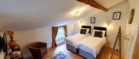 A bedroom with two beds, a chair, a small table with items, a window with curtains, and a wooden ceiling beam.