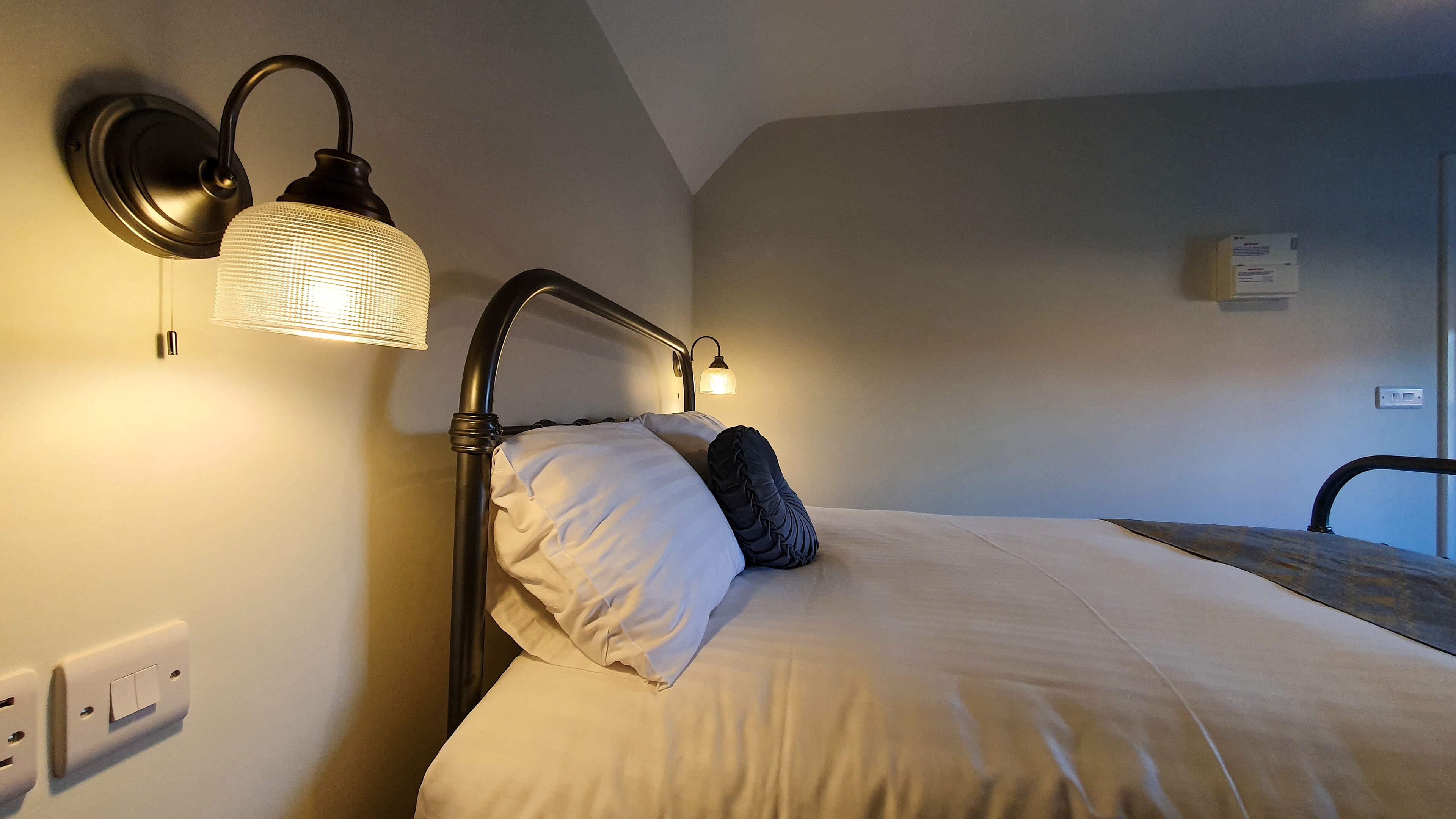 A neatly made bed with white linens and a dark pillow, a wall-mounted lamp, and a power outlet on the left side.