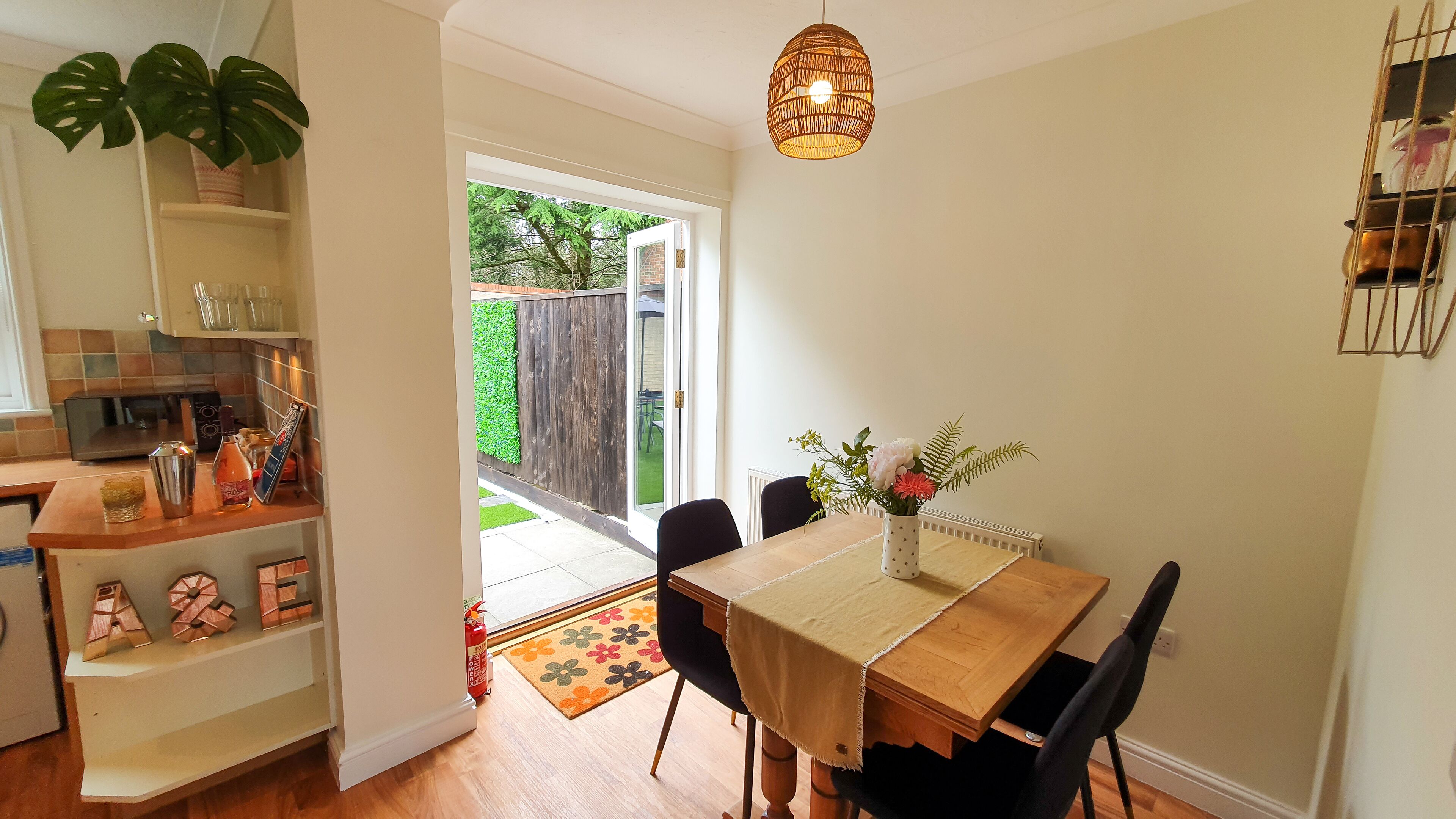 A dining area with a table, chairs, and a vase of flowers. There is a kitchen area with a plant and a shelf with various items.