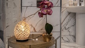 Une salle de bain avec un mur en marbre, un lavabo, un miroir, une lampe décorative et un pot d’orchidée.