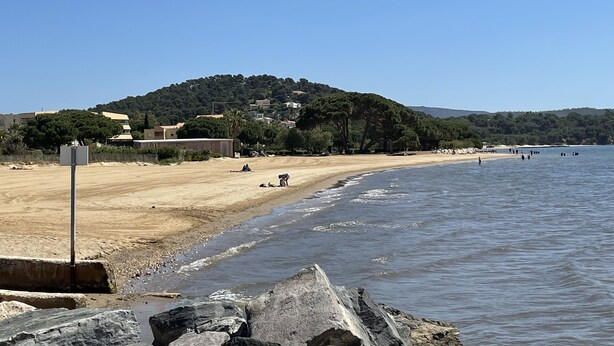 Ein Strand mit sandigem Ufer, einem steinernen Mole und einer Hügelkette mit Gebäuden im Hintergrund.
