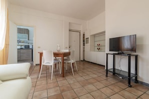 A living room with a TV on a stand, a dining table with chairs, a sofa, and a kitchen visible through an open door.