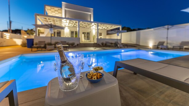 Zona de piscina moderna con una botella de vidrio transparente y aperitivos en una mesa, con vista a un edificio contemporáneo y una piscina.