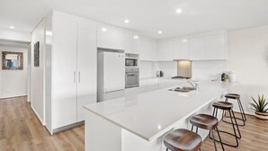 A modern kitchen with white cabinetry, a central island with stools, and a built-in oven. - Villa Serene 3, 6-10 Market Street -  Surfside Apartments WiFi, water views, Air Con,  Pool (Fingal Bay)
