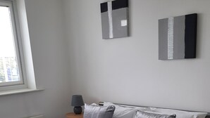 Een netjes opgemaakt bed met witte lakens en grijze kussens, een nachtkastje met een lamp, en twee abstracte schilderijen aan de muur.