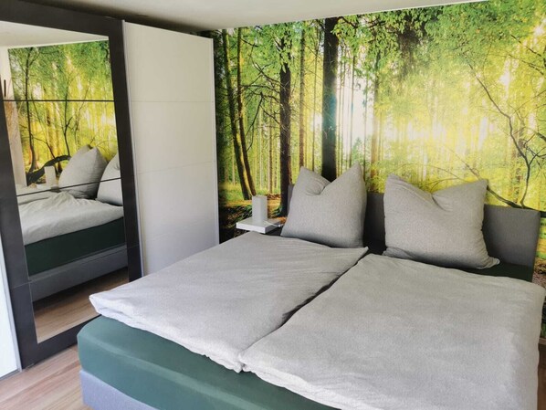A modern bedroom with a large bed, a nightstand, and a forest-themed wall mural. - Ferienwohnung Harnisch (Aue-Bad Schlema)