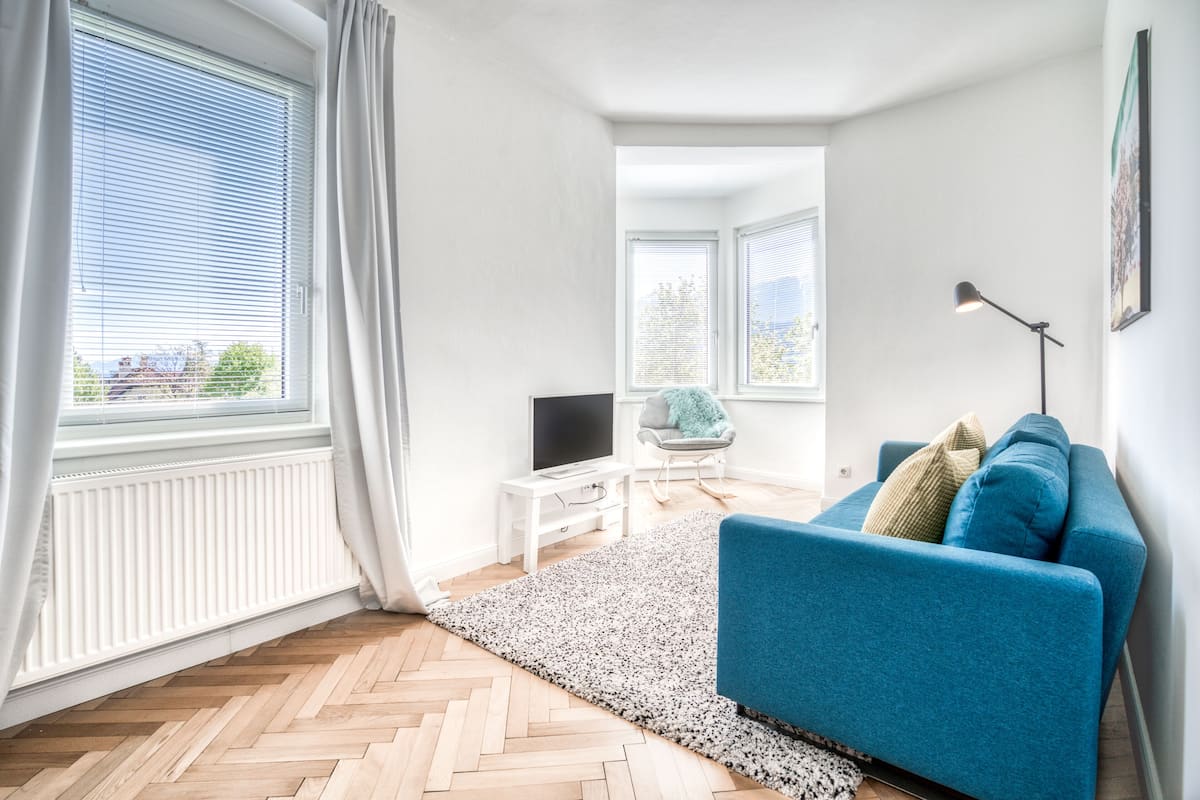 Una sala de estar moderna con un sofá azul, un televisor de pantalla plana, una pequeña mesa blanca y un ventanal con cortinas.