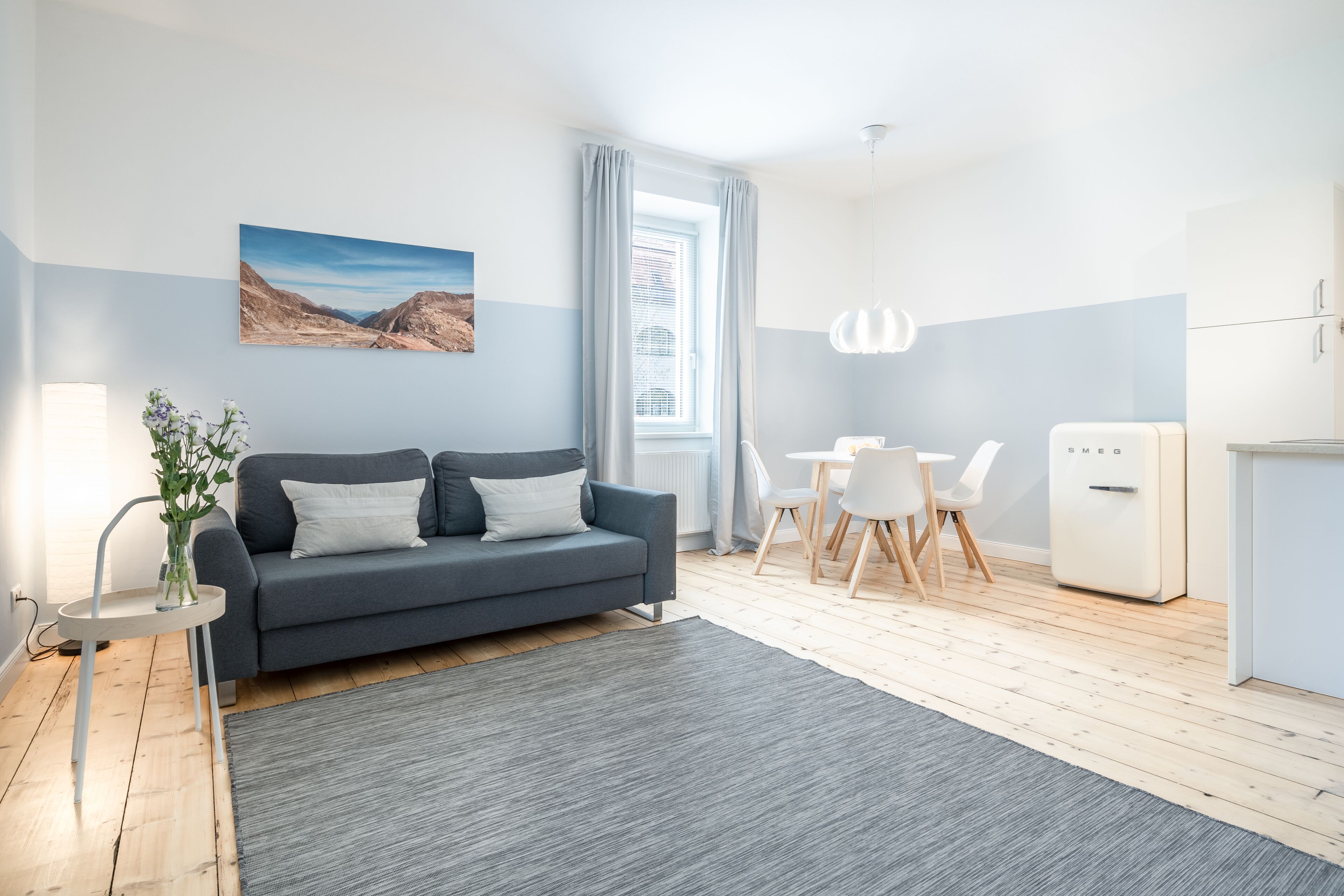 A modern living room with a grey sofa, a dining table with chairs, a small white fridge, and a window with curtains.