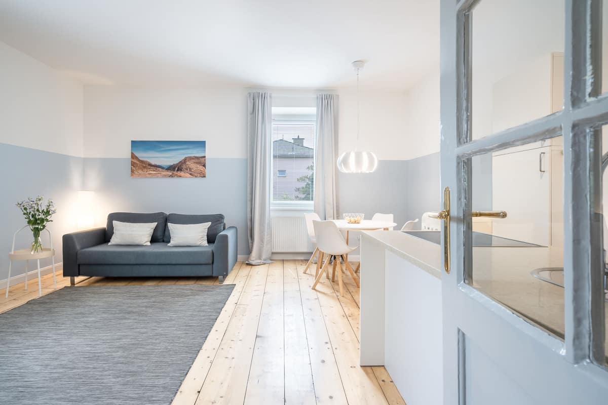 Una sala moderna con un sofá gris, un comedor con mesa y sillas, y una cocina con mostrador y armarios.