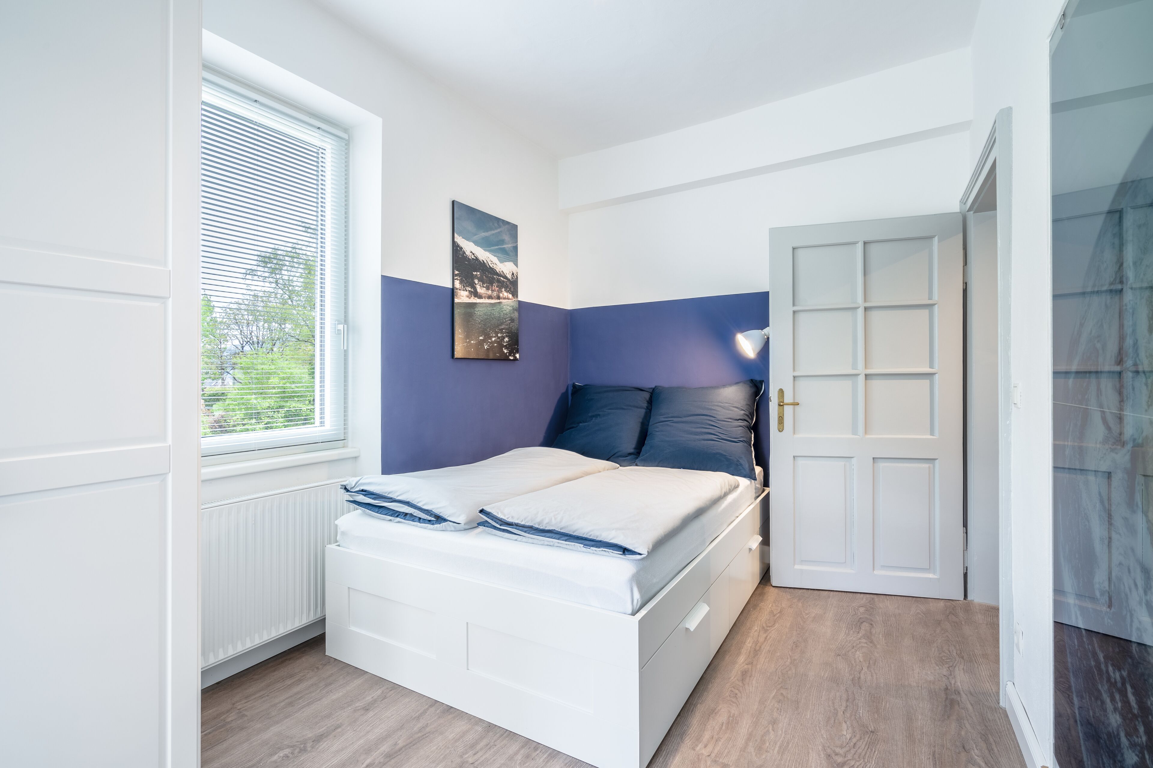 Ein Schlafzimmer mit einem Bett, einem Fenster mit Jalousien, einer Tür und einem Wandbild.