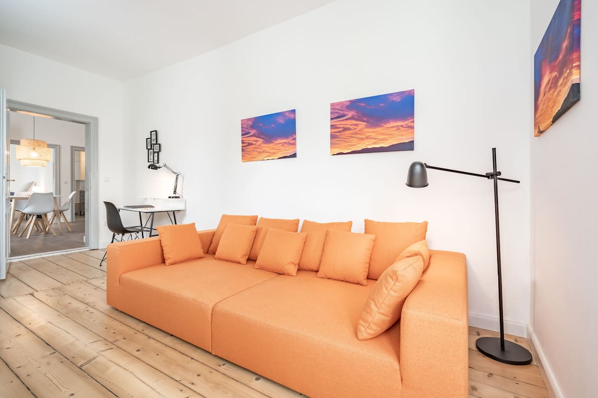 Una sala moderna con un sofá grande color naranja, una lámpara de pie y dos cuadros con motivos de atardecer en la pared.