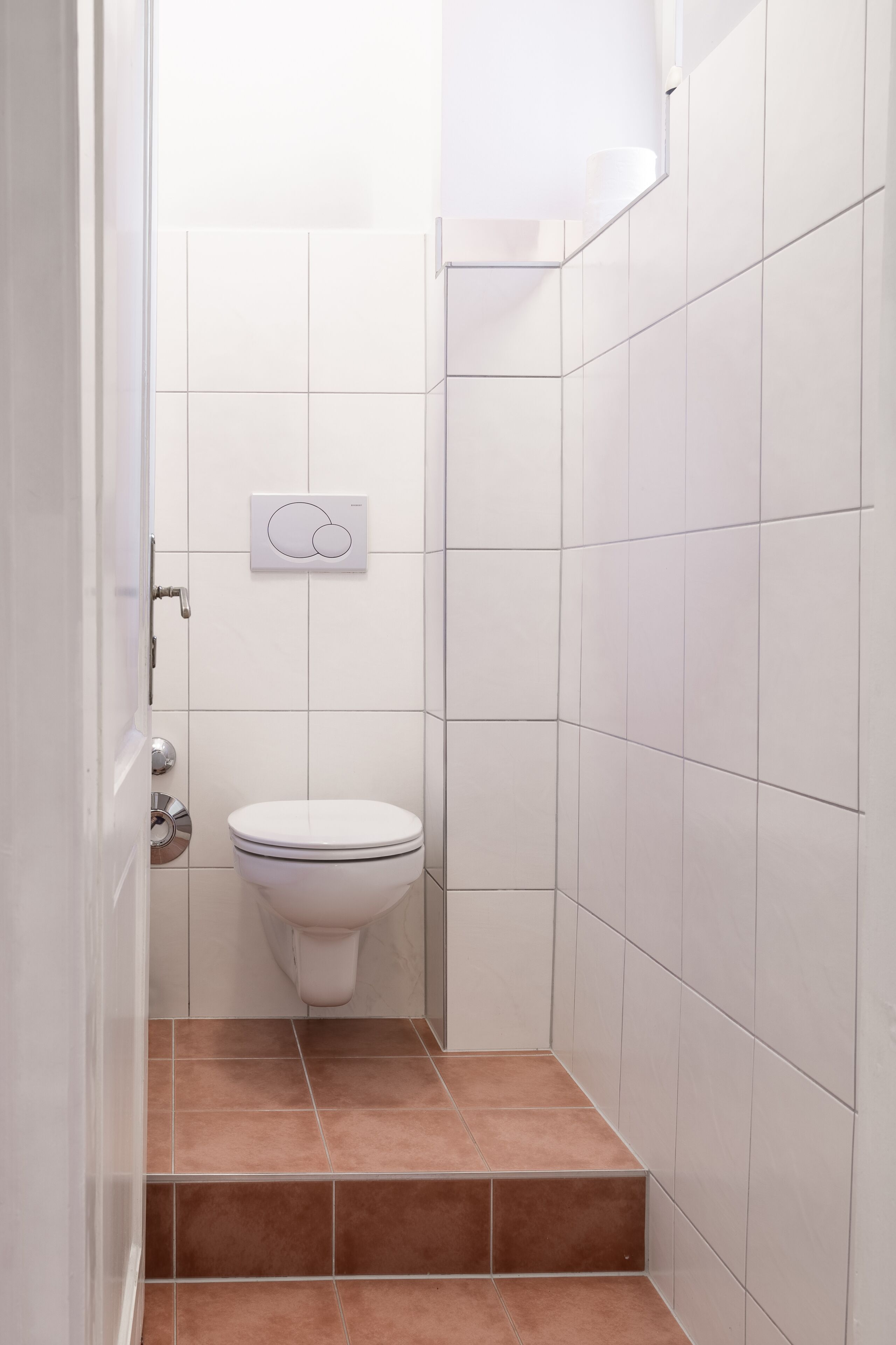 A small, white-tiled bathroom with a toilet and a flush handle.