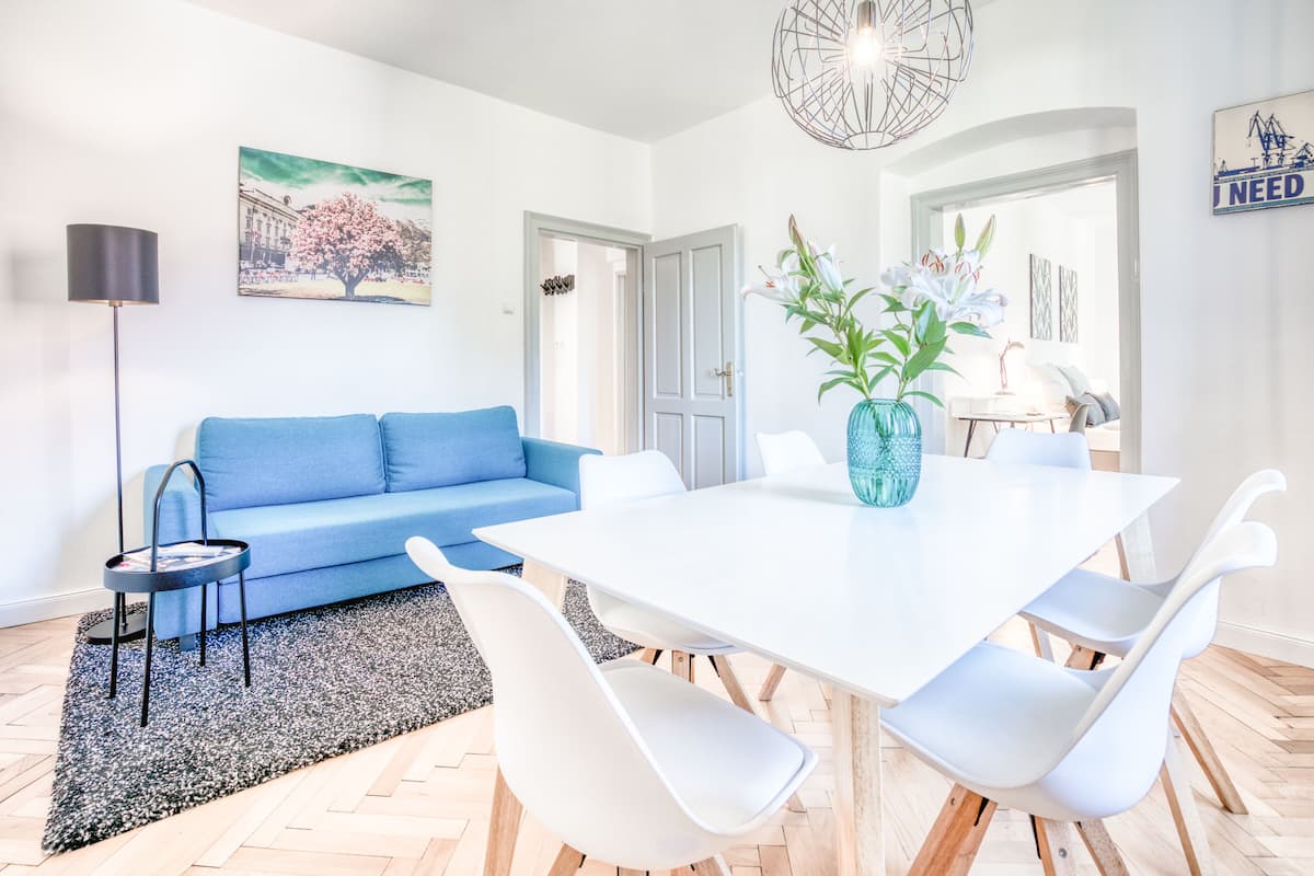 Un salón moderno con un sofá azul, una mesa de comedor blanca y sillas, una lámpara de pie negra y un jarrón con flores.
