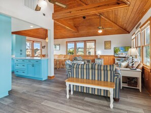A spacious living area with a wooden ceiling, a blue kitchen, and a large window overlooking the outdoors. - Waddle Inn: 255ft to Beach, 1-Min Walk, Duck OBX (Duck)