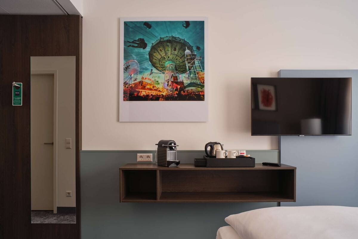 A hotel room with a bed, a TV mounted on the wall, a painting of a carnival scene, a coffee maker, and a small shelf with a kettle and mugs.