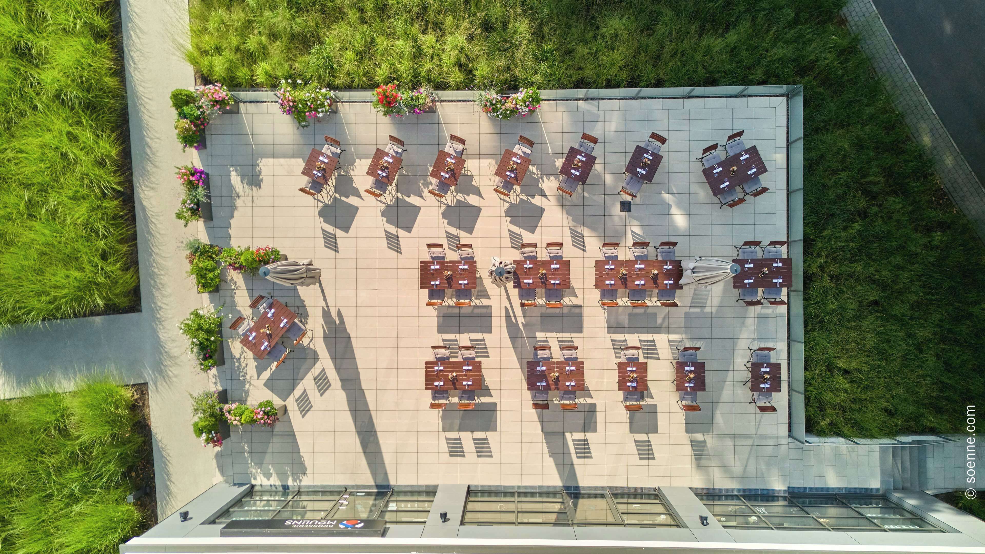 An aerial view of a rooftop patio with multiple tables and chairs arranged in a grid pattern, surrounded by lush greenery and flower planters.