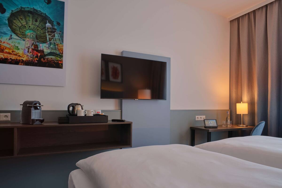 A hotel room with a bed, a desk with a coffee maker, a TV, and a painting on the wall.