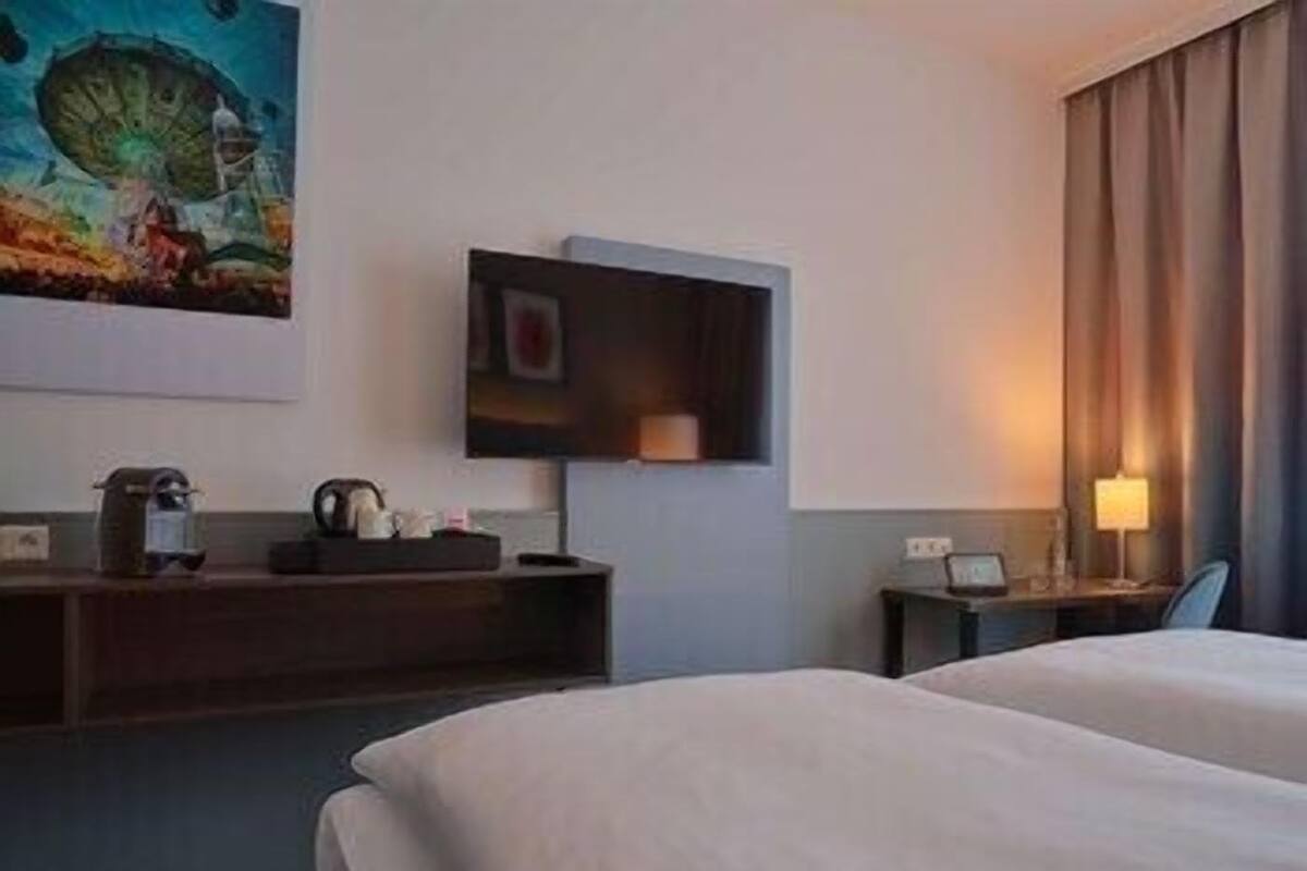 A hotel room with a bed, a TV mounted on the wall, a bedside table with a lamp, and a painting on the wall.